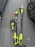All three tools laid out on driveway between two vehicles: compact hedge trimmer, long-reach hedge trimmer, corded pole chainsaw