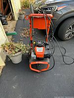 Front view of an orange Husqvarna pressure washer with hoses and spray gun sitting on a blacktop driveway next to a plant and parked cars.