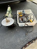 Green scroll saw with round metal table on left and clear plastic bin with various handheld power tools and batteries on the right, placed on concrete ground.