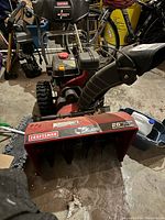 Angled front view showing red auger housing, chute assembly, control panel and drive tires