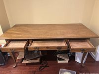 Photo showing the front view of the solid oak table with all three drawers open, visible ornate metal hardware, carved legs, and wooden floor beneath.