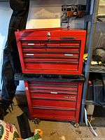 Photo of a red Task Force metal tool chest composed of two stacked units with drawers and a central lock in the top unit. Located in a garage or workshop environment with various tools and equipment nearby.