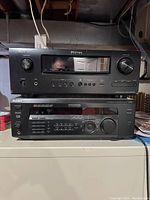Two stacked black audio amplifiers: Denon AVR-1600 on top and Sony STR-D565 below on a white surface against a basement background. Both units show their control panels with buttons, knobs, and displays visible.