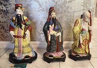 Photo of three Chinese porcelain statues depicting fortune, rank, and longevity, placed on dark wooden stands against a light background.