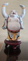 Front view of white porcelain Buddha sculpture with raised hands and colorful robe on dark wooden stand.