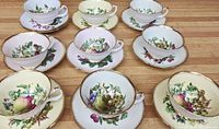 Nine bone china cups and nine matching saucers arranged in rows on wooden surface, each decorated with different fruit designs and gold trim