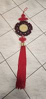 Photo of a Chinese hanging sign on a tiled floor. The sign includes a circular carved dark wood frame with a round cream-colored stone or ceramic piece in the center, red Chinese knotting above and below the wood frame, and long red tassels hanging down.