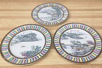 Three Chinese porcelain plates placed on a wooden surface, showing colorful rim patterns and detailed mountain scenery paintings on the plates' face.
