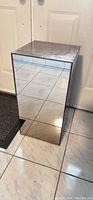 Full view of glass mirror rectangular stand placed on tiled floor against door. Shows reflective mirrored surfaces on all sides and top, revealing tile flooring reflections.