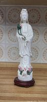 White porcelain Lady Buddha statue holding a flower, standing on wooden stand, detailed view showing delicate craftsmanship and floral accents.