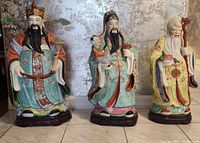 Photo showing three large, colorful porcelain statues known as the '3 guys' representing fortune, rank, and longevity, each seated on a carved wooden stand.