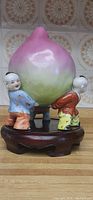 Front view of porcelain peach sculpture held by three small sons, displayed on a carved wooden stand with tiled background.