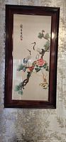 Full view of framed embroidered textile art showing two cranes on a pine tree branch with flowers in a brown wooden frame hanging on wall.