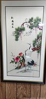Vertical silk picture showing two cranes near evergreen tree and flowers, framed in a black frame