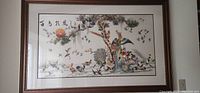 Framed silk embroidered picture showing various birds in colors around a tree, with Chinese characters on top left, glass covered and dark wood frame.