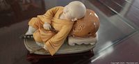 Photo showing small stone sculpture of a boy sleeping on a rounded bed with patterned pillow, placed on a dark carved wooden stand, showcasing details of the figure and fabric folds.