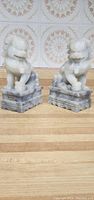 Photo showing two white stone lion bookends with detailed carving and rectangular bases on wooden surface in front of patterned tiled wall.