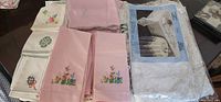 Photo showing various folded linens and napkins, mostly pink with floral embroidery, and white or cream linens with geometric and floral embroidery. Also shows a packaged lace tablecloth in its original wrapping.