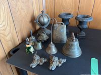Overview of all items including various antique bells made of brass, cast iron and glass, two miniature cast iron urns, and a pair of decorative cast iron feet placed on a black surface against wood paneling.