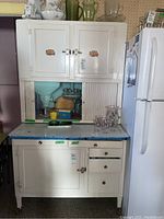 Front view of white antique Hoosier cabinet showing upper cupboards, lower cupboards and drawers, with blue countertop, adjacent to white refrigerator.