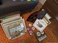 Overview of multiple framed paintings, prints, decorative fan, and smaller framed artworks arranged on the floor