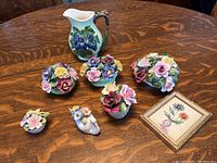 Photo showing group of bone china floral figurines and white pottery pitcher with grapevine motif, plus small framed flower art.
