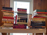 Stacked collection of multiple books of varying sizes and colors on a shelf beneath a window, showing a wide variety of titles including cookbooks, history, and novels.
