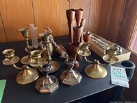 Photo showing brass, silver plate, and wooden candle holders along with brass bells and accessories, arranged on a black surface in front of a wood-paneled wall.