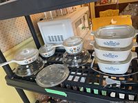 Three Corning Ware casserole dishes with lids, three small Corning Ware pots with lids, two glass bowls shown on a black shelf in a kitchen setting.