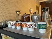 Overall view of two coffee pots, six ceramic Royal Alma mugs with colorful patterns, and six rocks glasses (five regular size and one smaller).