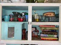 Two upper cupboards showing assorted glassware including juice glasses, glass candy dishes, a vintage hand mixer, salt and pepper shakers, and cookbooks.