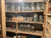 Wooden shelves in basement filled with various sizes of clear glass mason jars with metal lids and some empty jars, plus clay pots on lower shelf.