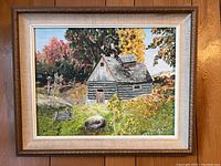 Framed oil painting on canvas panel showing a colorful autumn landscape with a log cabin. The frame is gold with a beige inner trim.