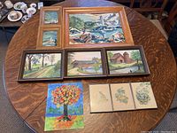 Overview photo of all ten artworks arranged on table showing prints and paintings with frames.