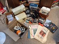 Full lot including radios, cassette recorder, Grundig emergency radio, Downton Abbey DVD set, CDs, cassettes, sheet music, power strips and adapters on carpeted floor.