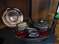 Silver plated teapot, sugar container, creamer placed on an oval wooden tray with silver rim handles, with round decorative silver plated tray beside.