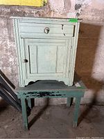 Wood cabinet painted pale green with drawer and door showing wear, metal ring pull on drawer and wood knob on door, placed on worn green wooden table with peeling paint.