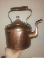 Front angled view of the antique copper kettle showing overall shape, spout, handle with wooden grip, and lid finial.