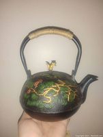 Side angled view of the cast iron teapot showing the handle wrapped in natural fiber and the gold and green embossed decoration on black base.