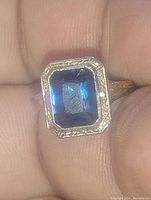 Close-up on finger showing emerald cut blue sapphire and engraved bezel.