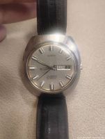 Front view of the Geneve men's wrist watch, showing dial with day and date at 3 o'clock, silver markers and hands, black leather strap.