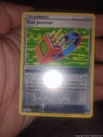 Close-up image of the Tool Jammer Pokémon Trainer card being held in hand. Shows holographic foil effect on the card surface and readable card text.