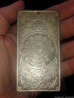 Rectangular metal bar held in hand showing side with Chinese zodiac animal wheel and symbols, surrounded by ornate engraved floral border.