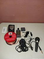 Photo showing handheld sound meter, walkman style radio, spool of speaker wire, two wired microphones with cables and adapter cords on white table.