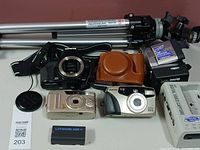 Photo showing Manfrotto tripod legs, Olympus camera body with strap, silver Samsung digital camera, Pentax Espio 105G film camera, battery and lens cap on white background