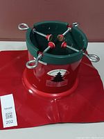 Red and green steel Christmas tree stand with four adjustable screws and plastic pads, showing label with Santa's Solution branding and a Christmas tree graphic.