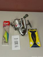Fishing reel and two fishing lures, one in box and one in packaging, all shown together