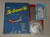 Photo showing 'From The Ground Up' blue aviation booklet with airplane illustration, APR flight computer in original red sleeve, and instruction pamphlet