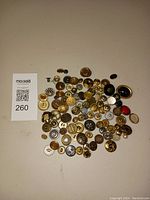 Lot of assorted metal and plastic sewing buttons laid out on a white surface with MaxSold QR code tag showing lot number 260.