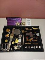 Overview of assorted men's accessories including two wristwatches, multiple keychains, cufflinks, a pen, belt buckle, tie clip and bow tie in display box.